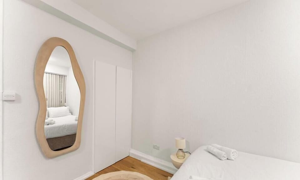 A wavy-edged wooden mirror leans against a white wall, reflecting a tidy bed in a minimalist, neutral-toned room. A simple wardrobe and small bedside lamp complete the clean, serene aesthetic.