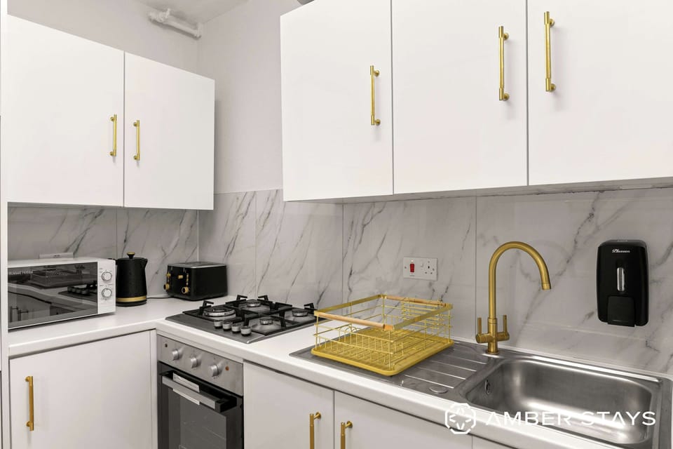 This modern kitchen features crisp white cabinetry accented by brushed gold hardware and a matching faucet. A marble-patterned backsplash ties the sleek, compact space together for a clean and elegant look.