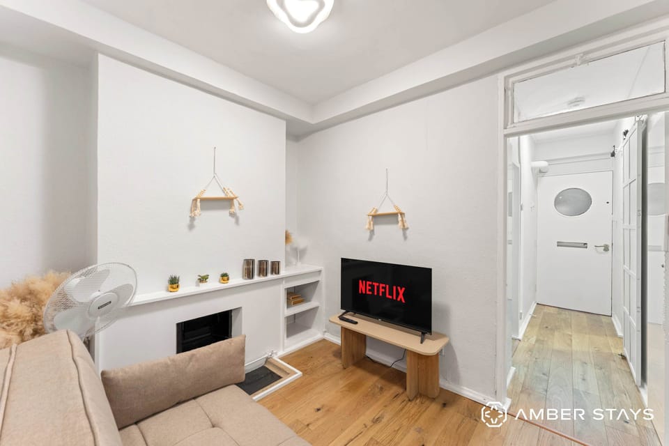 This minimalist living area features bright white walls, light wood flooring, and a neutral sofa tucked into the corner. A small TV sits on a wooden stand next to a built-in mantel decorated with subtle accents.