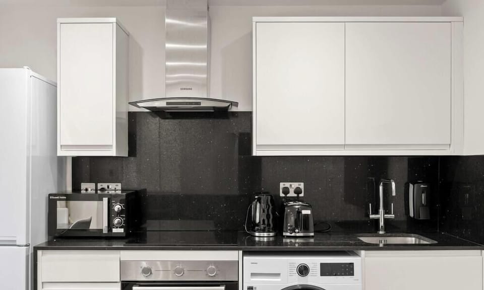 This modern, compact kitchen features minimalist white cabinetry contrasted by a dark, textured backsplash and black countertops. It is efficiently designed with integrated appliances, including a washing machine, refrigerator, and a sleek stainless 