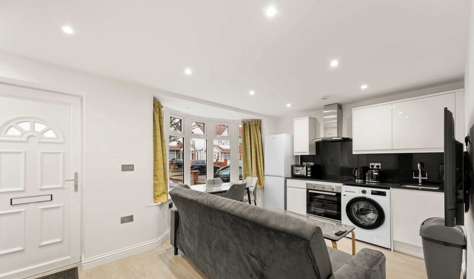 This compact living space features a modern white kitchenette and a grey sofa positioned near a bright bay window. Mustard-yellow curtains add a pop of color to the otherwise neutral, sunlit room.