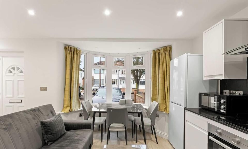 This bright, open-plan studio features a neutral seating area and dining set illuminated by a large bay window. Modern recessed lighting and a sleek, compact kitchen create an airy and efficient living space.