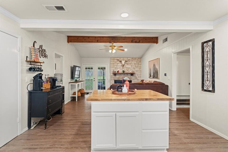 Wide-angle view of the open-plan living space from the kitchen island, showcasing exposed wood ceiling beams, a white kitchen island with butcher block top, coffee station, and a clear sightline to the stone fireplace and French doors beyond.