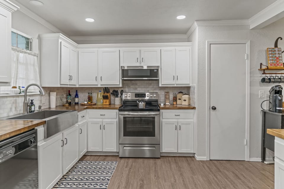 Crisp white shaker kitchen with butcher block countertops, a subway tile backsplash, stainless steel appliances, and a patterned runner rug — a bright, well-stocked space that balances farmhouse charm with everyday functionality.