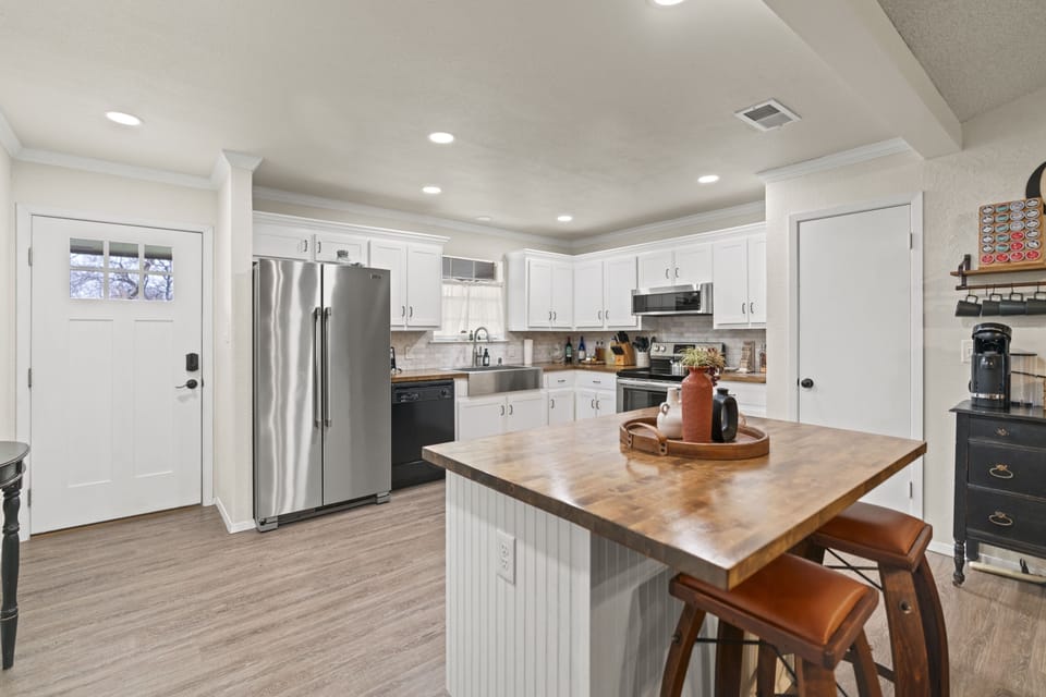 Open-plan layout viewed from the kitchen island, with a butcher block prep surface in the foreground, a coffee station tucked alongside dark cabinetry, and a clear view through to the fireplace and French doors — a connected, functional home base.