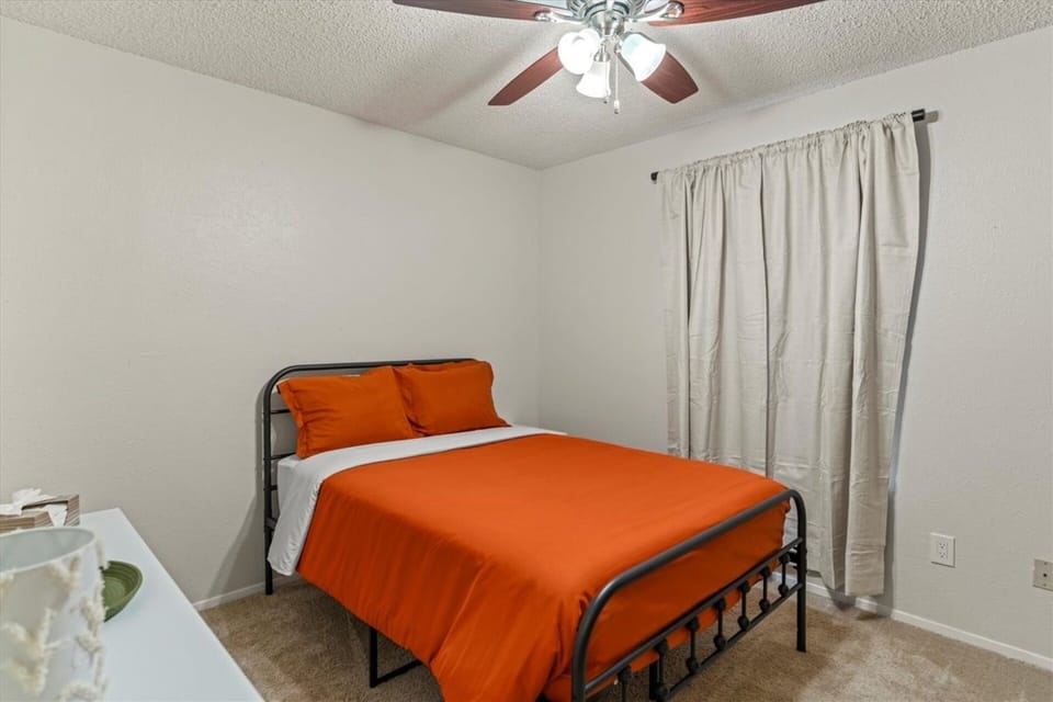 Simple and restful guest bedroom with a full-size black metal bed frame, bold orange bedding, neutral linen curtains, and a ceiling fan — a clean, no-fuss space that delivers comfortable, uninterrupted sleep.