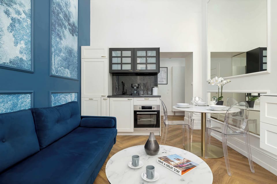 Another perspective of the living space highlighting the blue sofa and round coffee table. The kitchenette area with white cabinetry and dark upper cabinets is visible beyond.