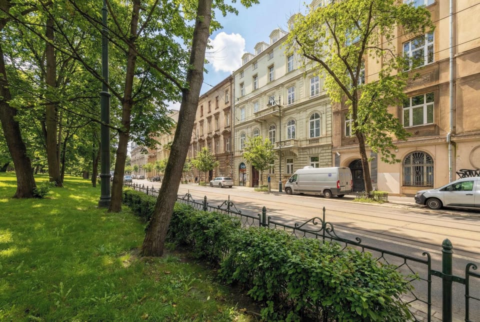 An exterior view of a tree-lined street with historic buildings featuring ornate facades. Green grass and mature trees frame the sidewalk. The atmosphere feels peaceful and residential.