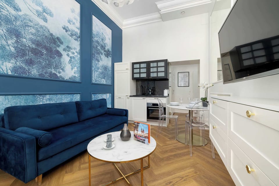 A stylish living area with a blue sofa positioned beneath oversized blue-toned wall art. A white dresser stands along one wall, and a round coffee table anchors the seating area. The open layout leads toward a compact kitchenette in the background.