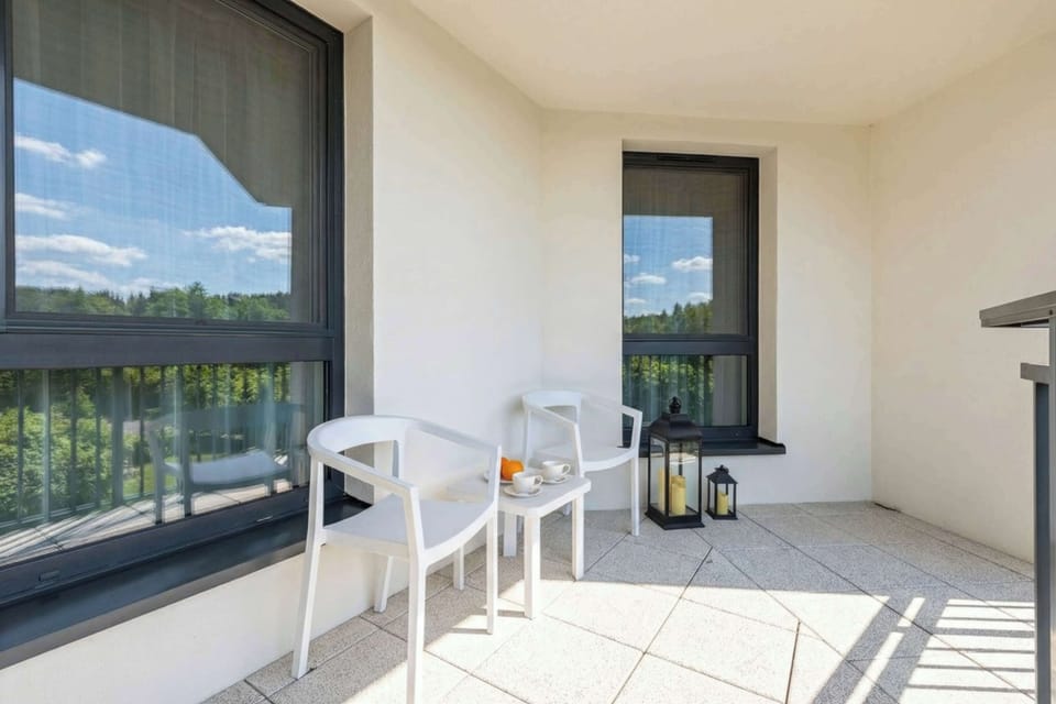 Two white chairs and a small table are set on the balcony, offering a quiet spot to enjoy the fresh air. The view overlooks the surrounding greenery and modern architecture of the estate.