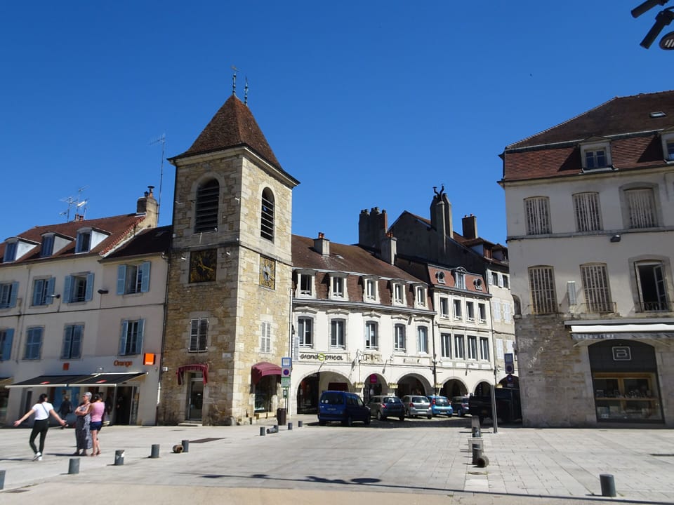 A charming town square showcases traditional Jura architecture, colourful façades, and a lively café atmosphere under clear blue skies.