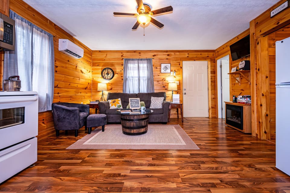 Living area with polished hardwood floors, gray sofa, matching armchair, barrel-style glass coffee table, and ceiling fan.