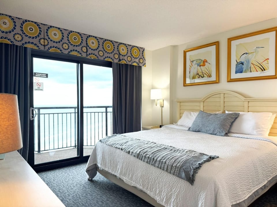 King bed with textured white bedding and blue-gray throw pillow faces sliding glass doors to oceanfront balcony.