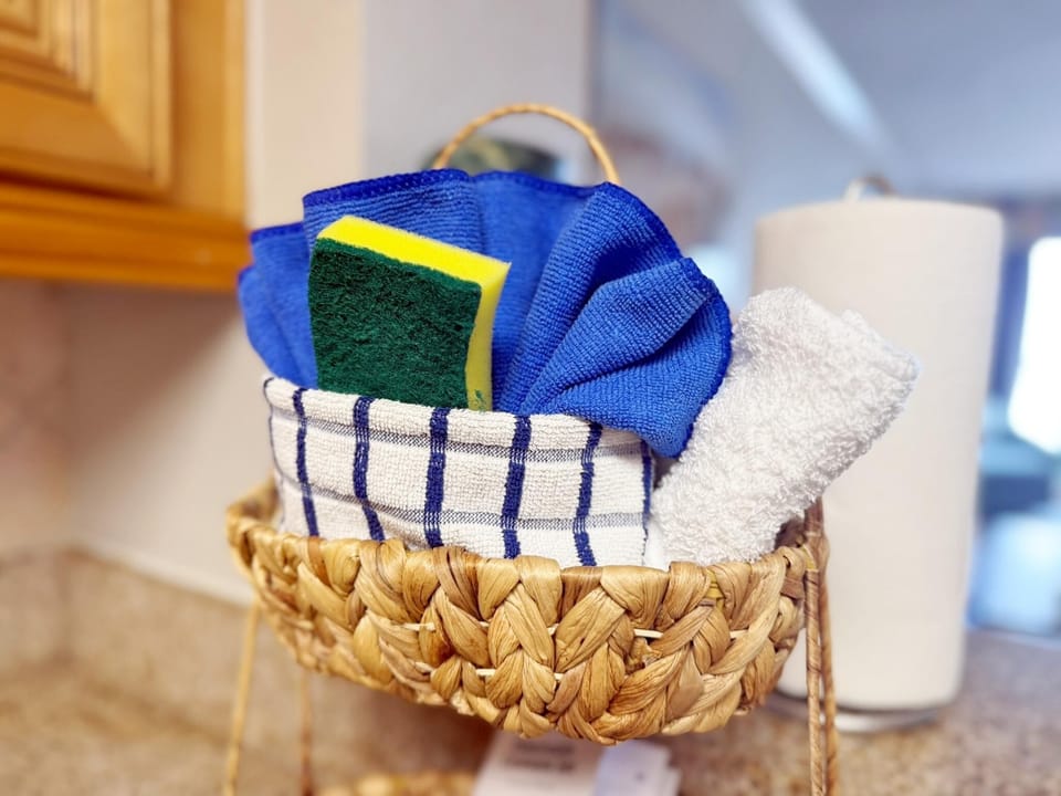 Woven basket holds blue microfiber cloths, a yellow and green sponge, and a striped kitchen towel.