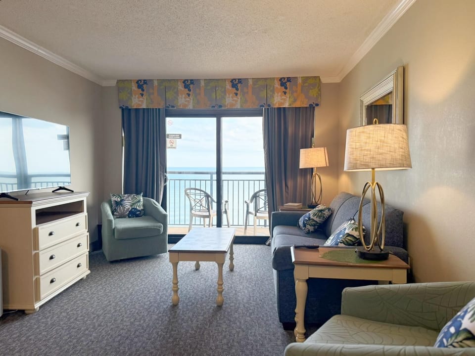 Living area with patterned carpet, floral valance, and sliding doors opening to an oceanfront balcony with two chairs.