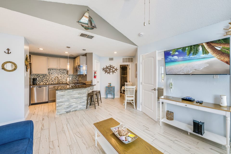 Relax in your coastal-themed living space featuring an open kitchen, nautical décor, and flat-screen TV with tropical beach views.