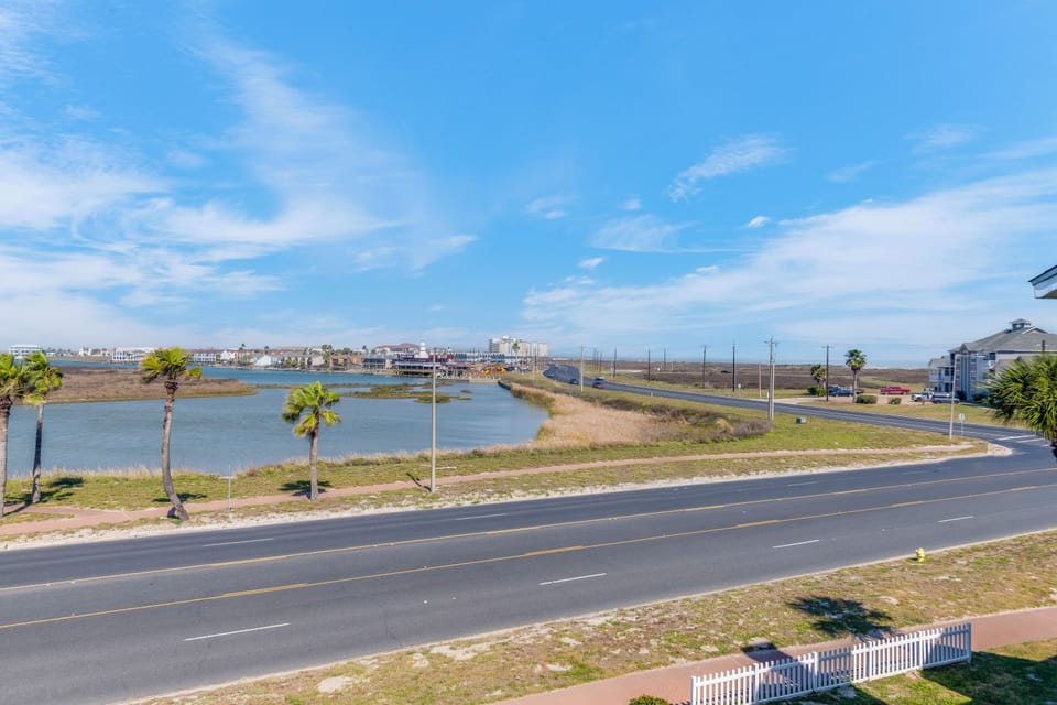 Peaceful coastal view featuring lagoon waters, palm trees, and distant beachfront buildings under bright blue skies.