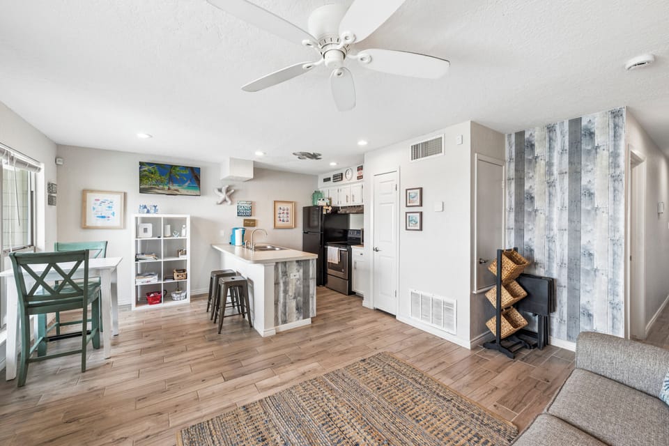 Your open-concept living space features modern coastal charm with comfortable seating, ceiling fan, and a fully equipped kitchen perfect for vacation meals.