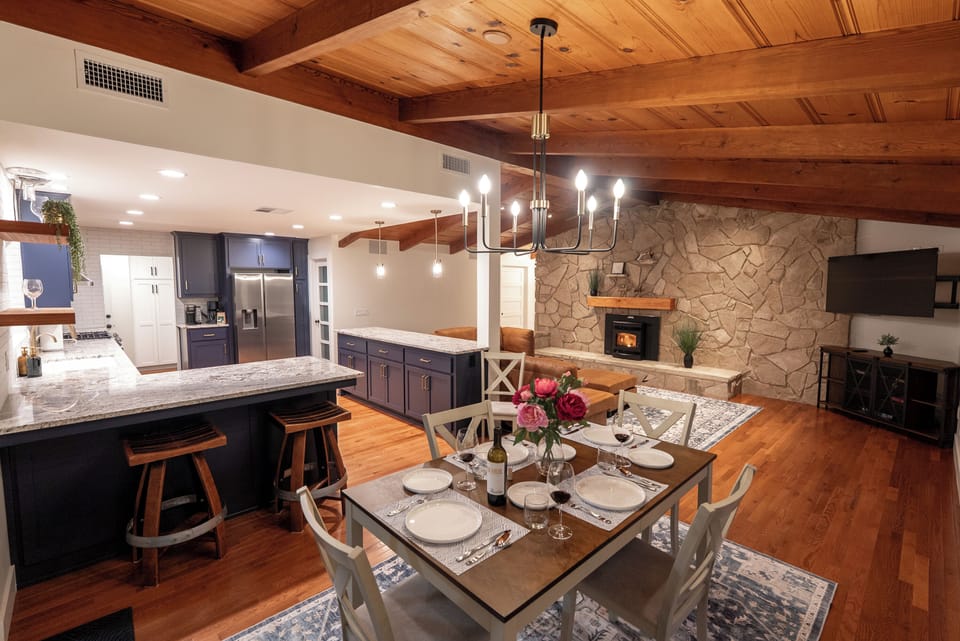 This open-concept interior features a warm wood-beamed ceiling, a floor-to-ceiling stone fireplace, and a set dining table illuminated by a contemporary chandelier.