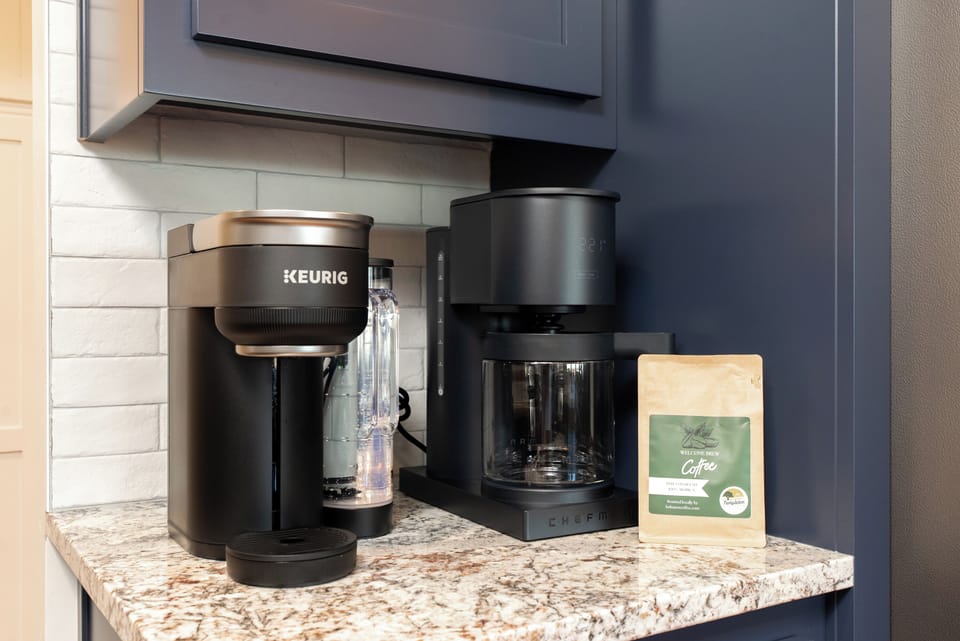 A dedicated coffee station featuring a modern Keurig pod machine and a Chefman drip brewer on a granite countertop, nestled against sleek navy cabinetry and a white subway tile backsplash.