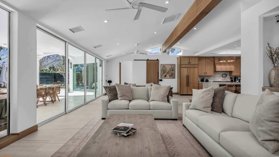 Bright open-plan living area with wood beam ceiling, floor-to-ceiling sliders, barn door, and kitchen opening to mountain-view patio
