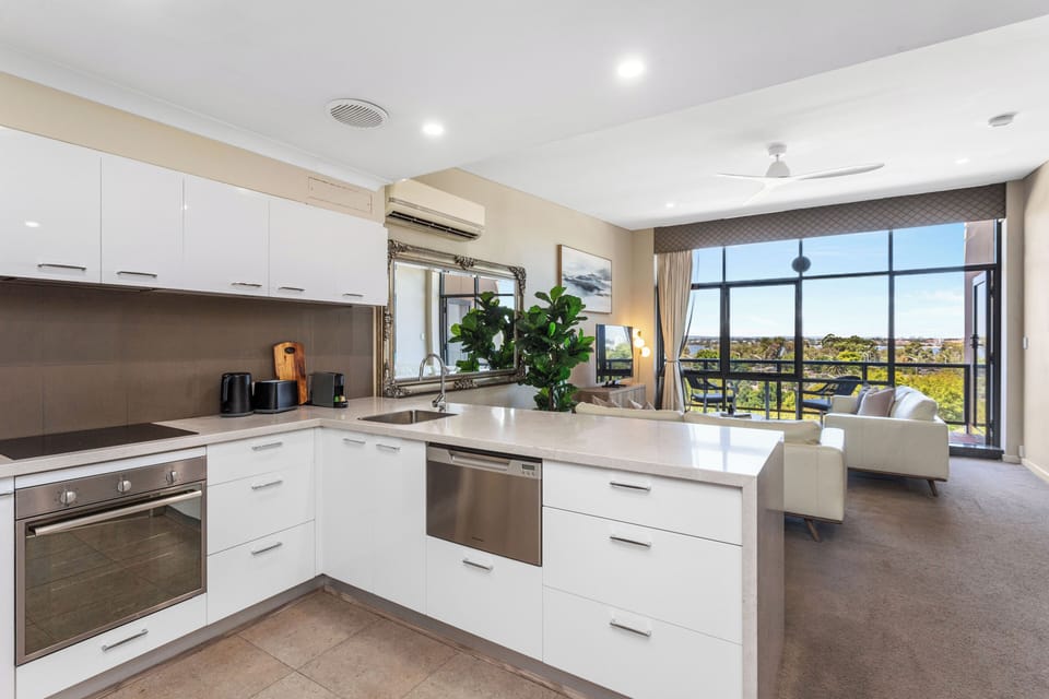 A bright, modern kitchen and living space featuring a stone waterfall island, sleek white cabinetry, and floor-to-ceiling windows that lead to a private balcony with scenic views.