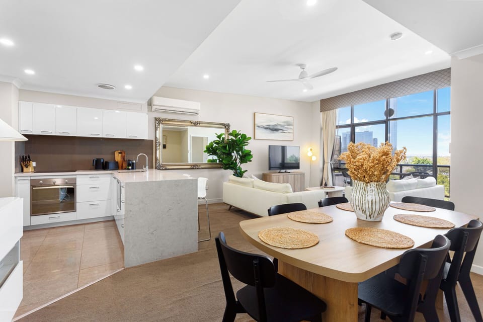 A bright, open-plan living and dining area featuring a modern kitchen with stone countertops and floor-to-ceiling windows that open onto a private balcony with scenic views.