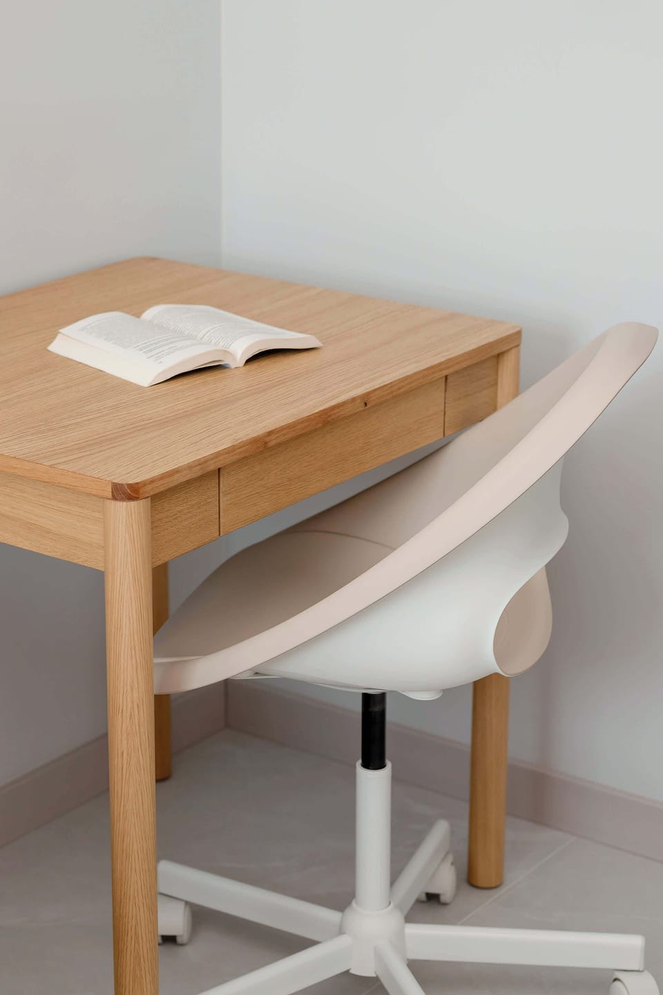 A focused shot of the workstation shows a minimalist wooden desk with an open book and a modern white swivel chair. The simple setup is located in a quiet corner of the bedroom.