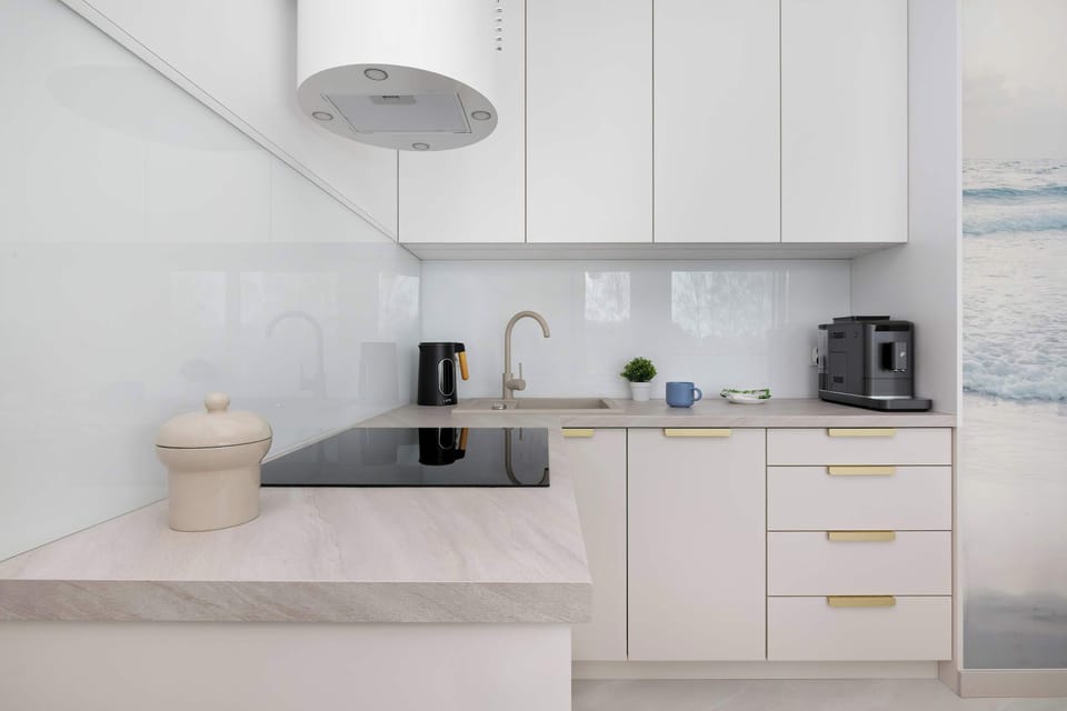 The compact kitchenette is designed with minimalist white cabinetry and a light wood-textured countertop. It includes a built-in induction hob, a coffee machine, and essential kitchenware for guest use.