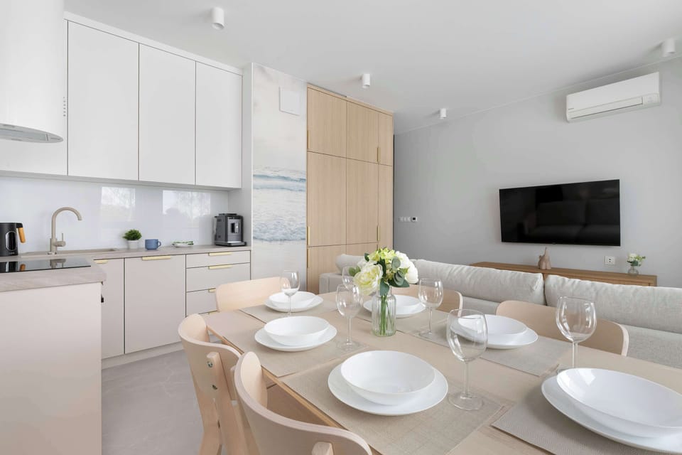 A functional dining space is set with white plates and wine glasses, ready for a group dinner. In the background, the kitchen and a wall-mounted TV are visible, showing the apartment's integrated layout.
