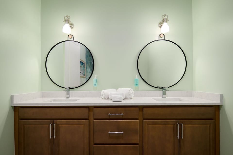 A serene green bathroom features modern round mirrors paired with warm wood cabinetry, exuding a calming coastal vibe.