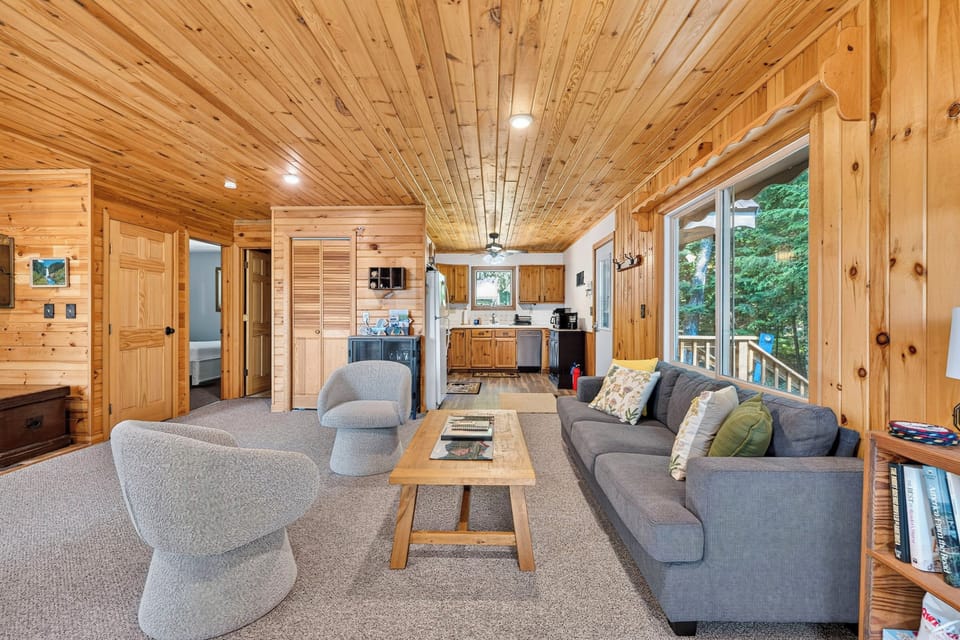 Large open living area with a gray couch, wood coffee table and two gray swivel chairs. in the back ground the small kitchen opens into the living area.