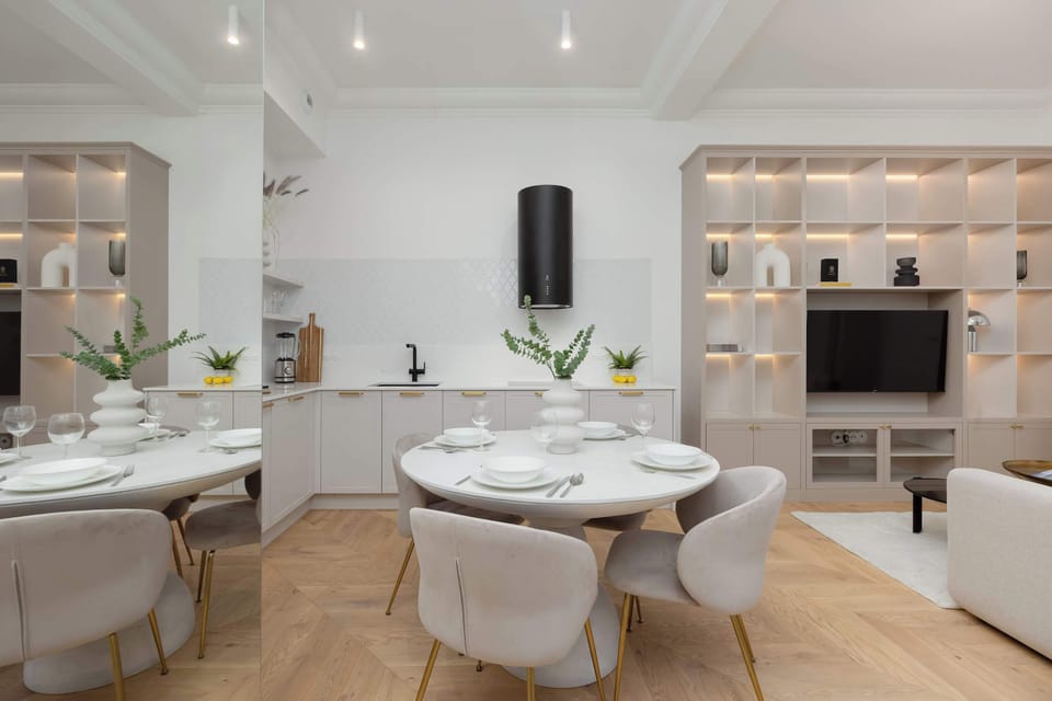 A dining table set with plates and glassware under warm ceiling lights. Shelving and kitchen cabinetry frame the area. The scene feels inviting and ready for guests.