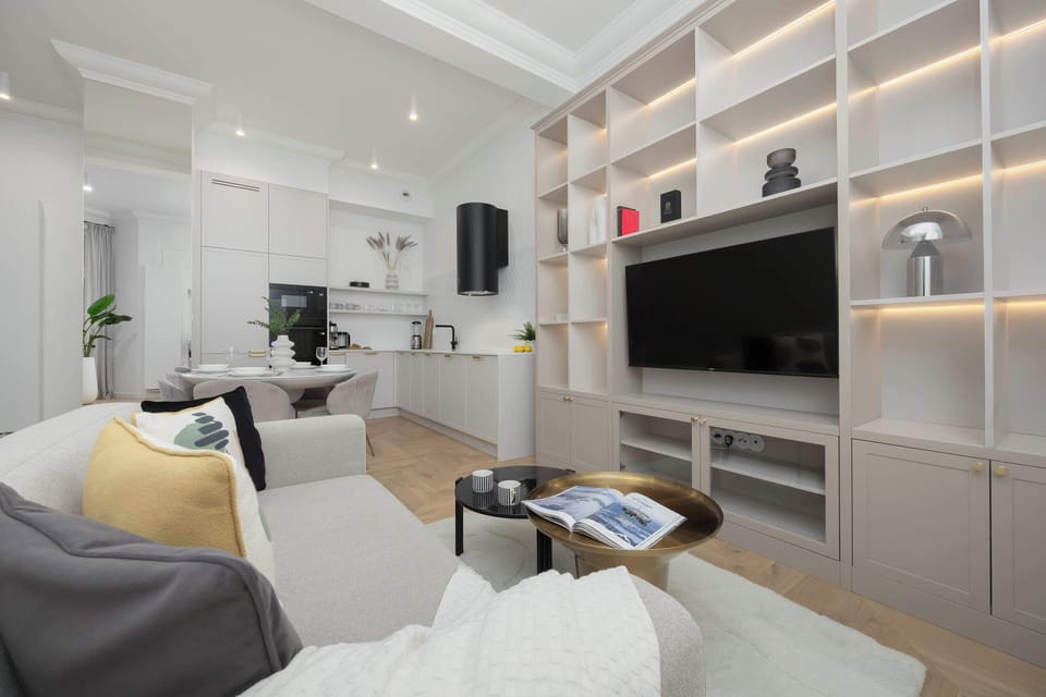 A living area featuring a sofa, armchair, and a wall-mounted TV surrounded by shelves. Neutral tones dominate the space. The layout feels comfortable and functional for daily living.
