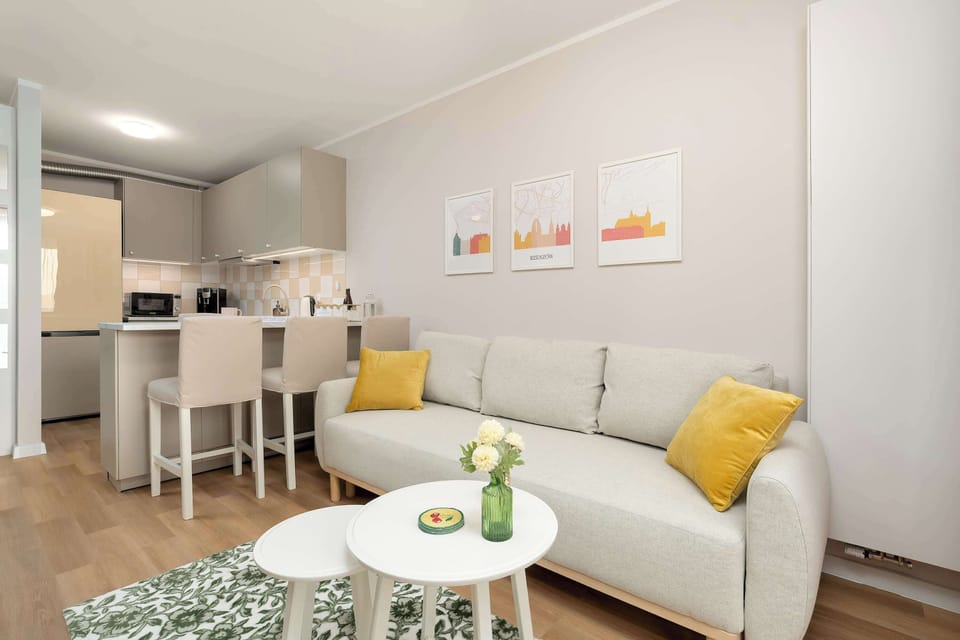A dining nook with a small table and chairs sits beside the living area. The sofa with yellow pillows appears again, reinforcing the apartment’s consistent design style.