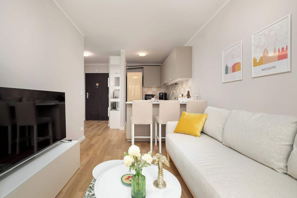 A long perspective of the living room looking toward the kitchen, showing the sofa, coffee table, and hallway beyond. Yellow pillows add warmth to the neutral palette.