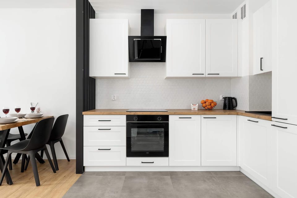 A bright kitchenette designed with clean white cabinetry, a black backsplash, and modern appliances. A wooden dining table with black chairs is partially visible, creating a seamless transition between cooking and dining areas.