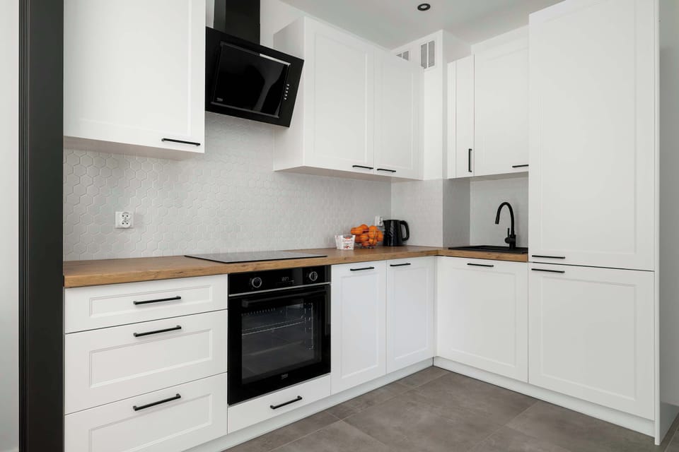 A modern L-shaped kitchen with white gloss cabinets, black handles, and an integrated black oven. The contrast between the light furniture and dark hardware gives the space a sophisticated look.
