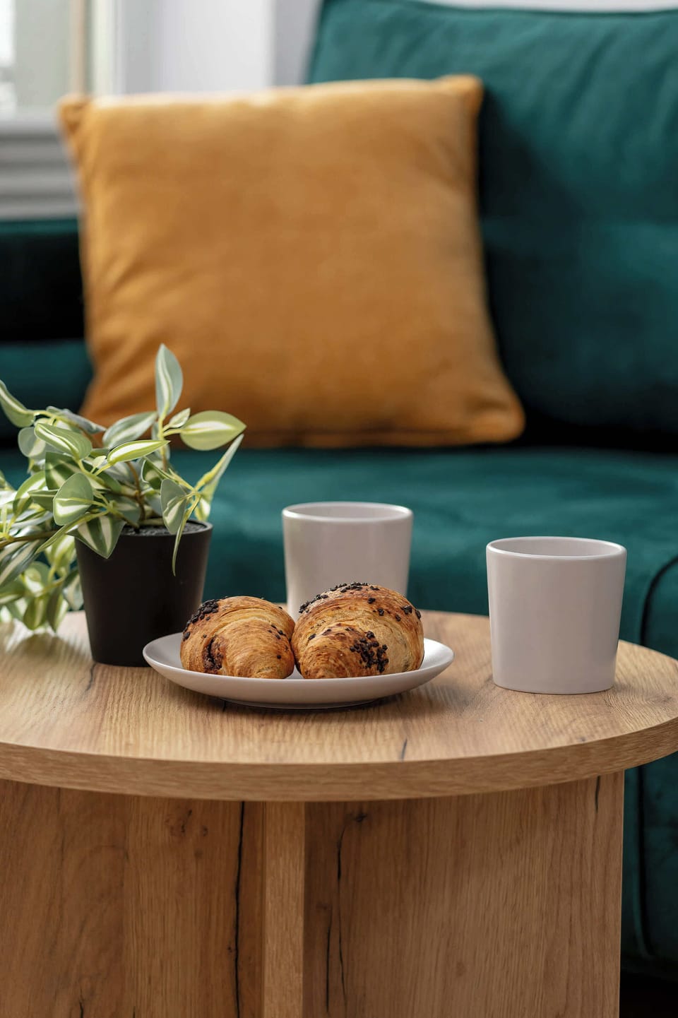 A lifestyle shot featuring a wooden coffee table set with fresh croissants and two mugs. The blurred green sofa in the background creates a warm, inviting "home away from home" feeling.