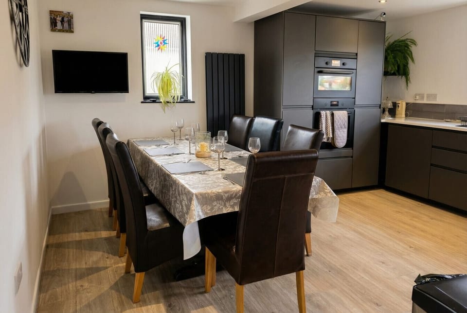 A dining table with set place settings and leather chairs occupies a room adjacent to dark grey fitted kitchen units, with a wall-mounted TV visible.