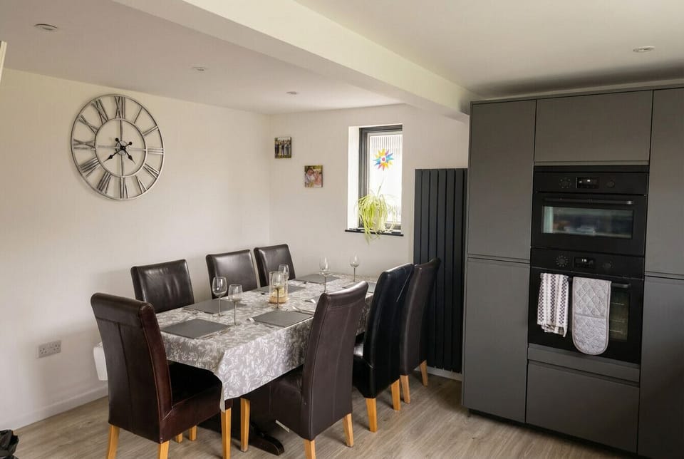 A dining table is set for a large-group meal alongside grey fitted kitchen units and a built-in double oven tower.