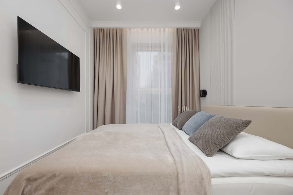 A primary bedroom view showcasing a large wall-mounted TV facing a plush bed with neutral-colored pillows and soft drapes.