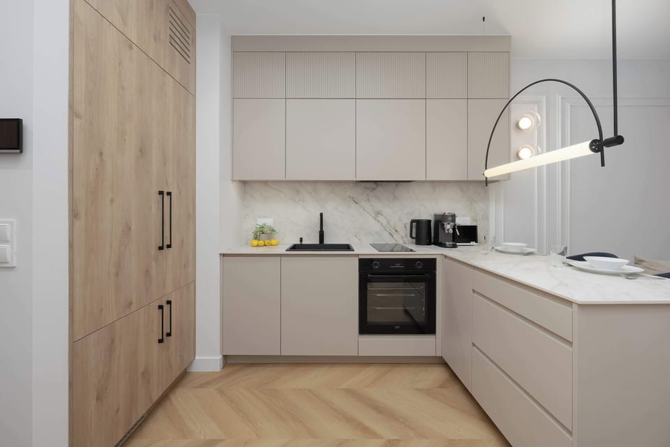 A sleek kitchenette layout showing light beige handleless cabinetry, a white marble backsplash, and light wood flooring throughout.