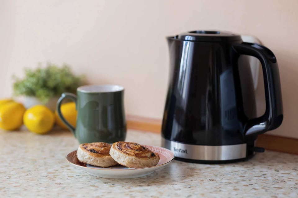 A close-up of a kitchen countertop with a black electric kettle, a cup, and some lemons. Wooden cabinets and a tiled backsplash form the background. The arrangement suggests a ready spot for making tea or coffee.