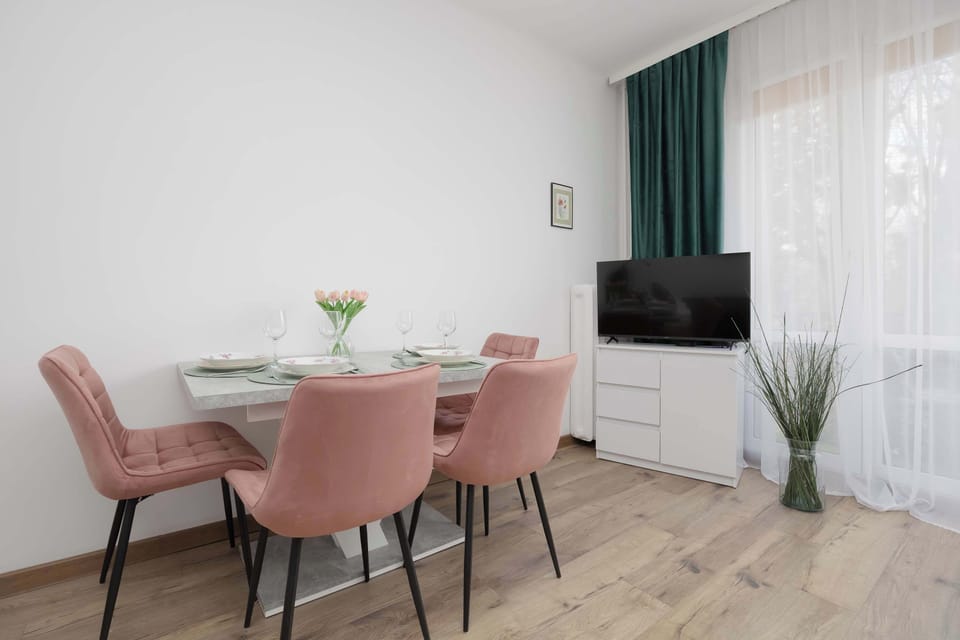 A dining area positioned near tall windows with sheer white curtains. Pink chairs surround a simple white table. Natural light brightens the space and highlights the light wood flooring.