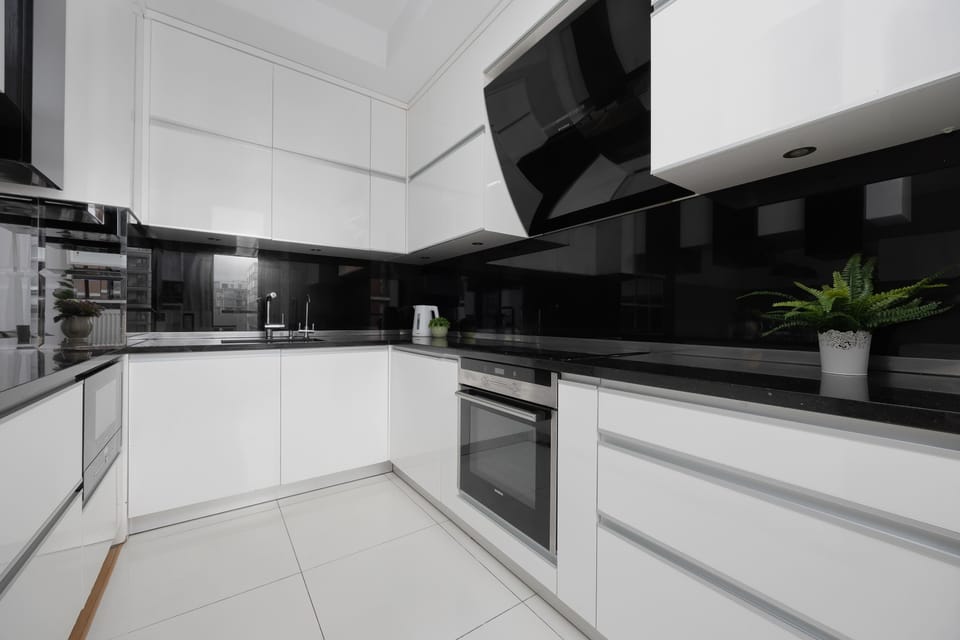 A contemporary kitchen showcases sleek white cabinetry contrasted with a black glossy backsplash and countertops. The L-shaped layout includes modern built-in appliances and bright tiled flooring.