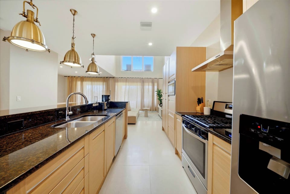 This bright and airy kitchen welcomes you with sleek granite countertops and warm wooden cabinetry. Pendant lights illuminate the workspace, while large windows bring in natural light and a cheerful view.