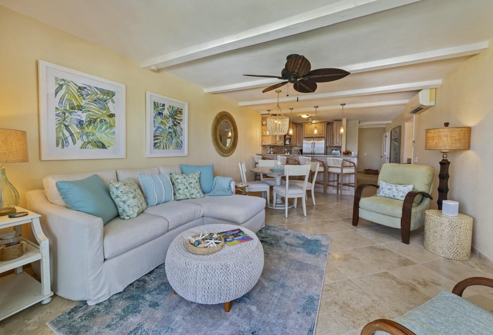 Open living area with sectional, woven round ottoman, and tropical leaf artwork.