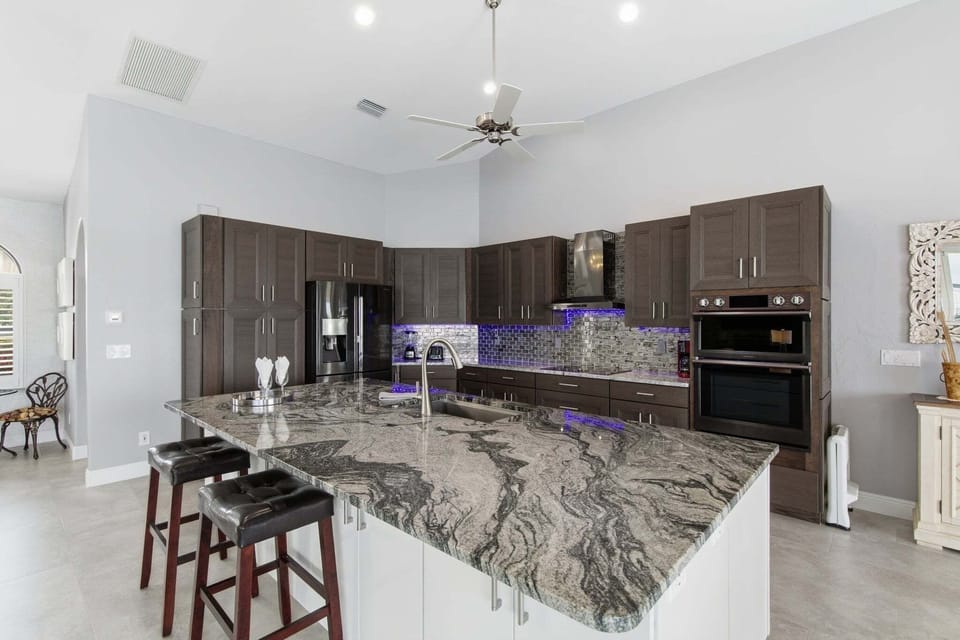 Gourmet kitchen featuring dark wood cabinetry, dramatic granite island with bar stools, double wall ovens, and LED backsplash lighting