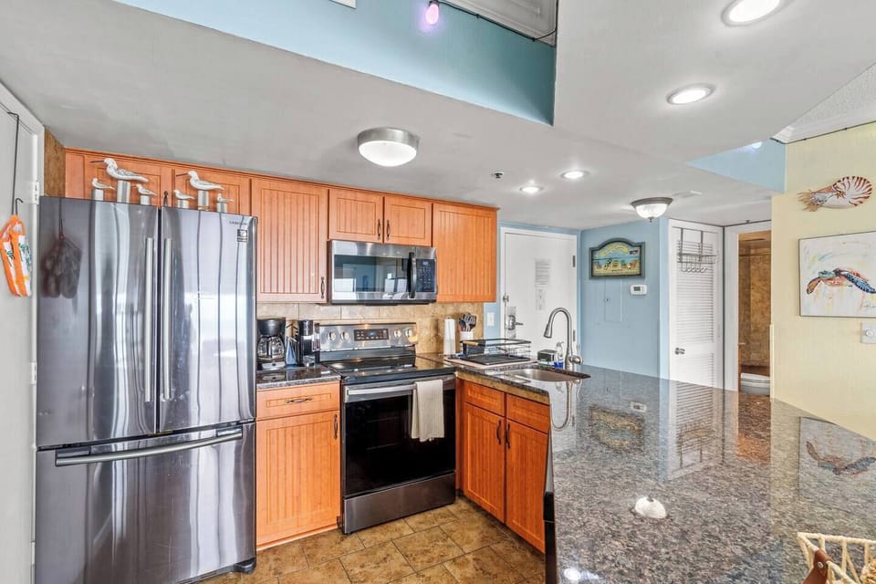 Fully equipped kitchen with stainless steel appliances, including refrigerator, stove, oven, microwave, dishwasher, sink, Keurig, drip coffee maker, and breakfast bar seating for three.